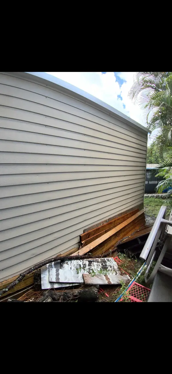 The side of a building with weathered siding and boards stacked against the base.