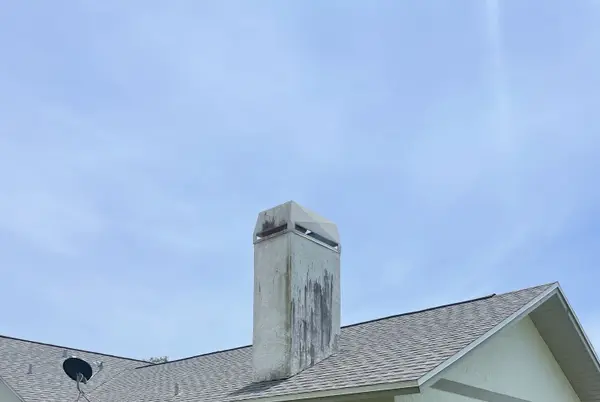 A stained chimney and shingled roof slope against a clear sky.