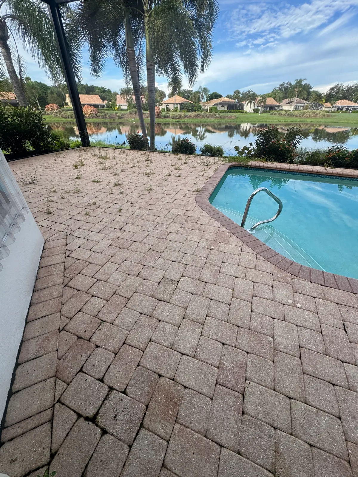 A paver pool deck overgrown with weeds, with a pool and lake beyond.