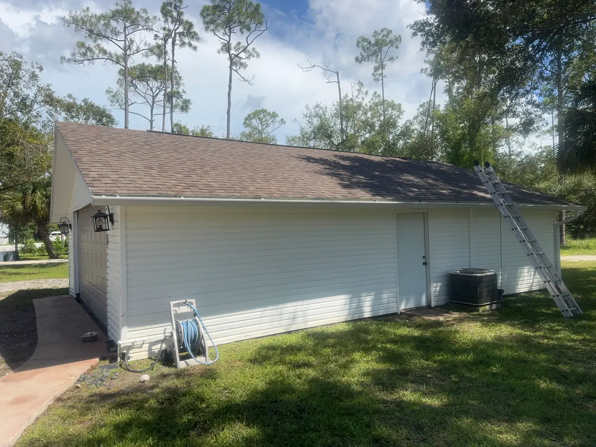 A white garage wall with a brown shingled roof, a hose reel and an extension ladder.