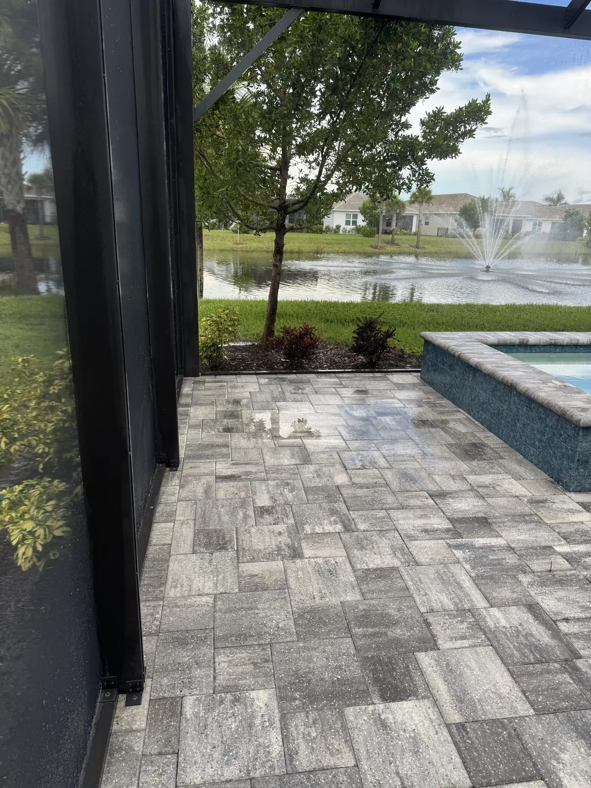 A paver patio beside a pool with a fountain beyond, seen through a screen enclosure.